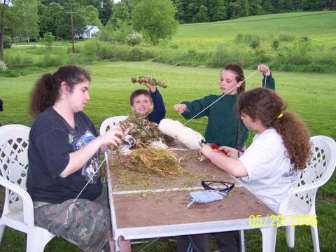 Family sitting at a table outdoors wrapping cedar leaves with yarn .